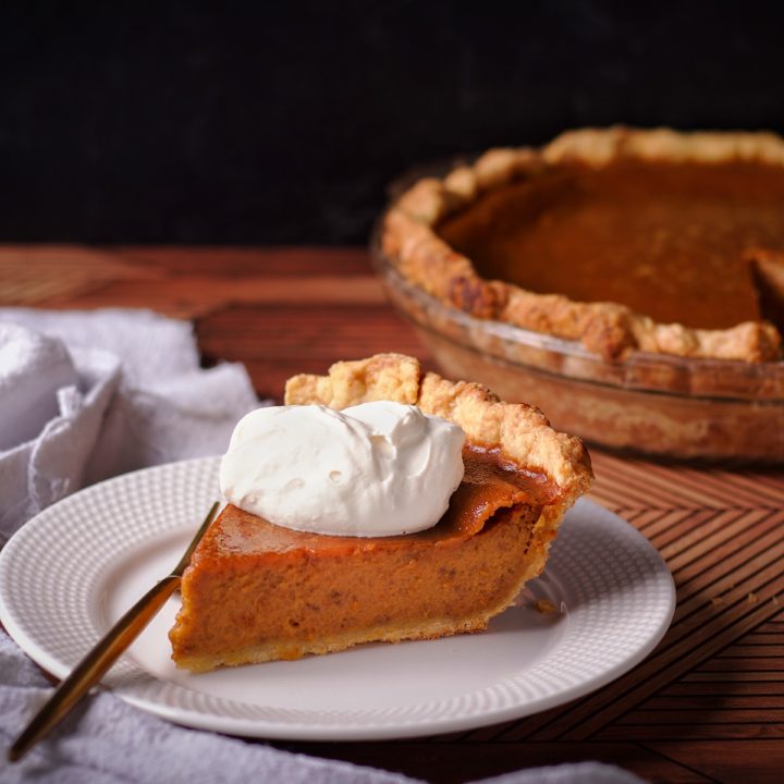 A slice of Maple Pumpkin Pie with Maple Whipped Cream on a plate with a fork next to it and the rest of the pumpkin pie in the background.