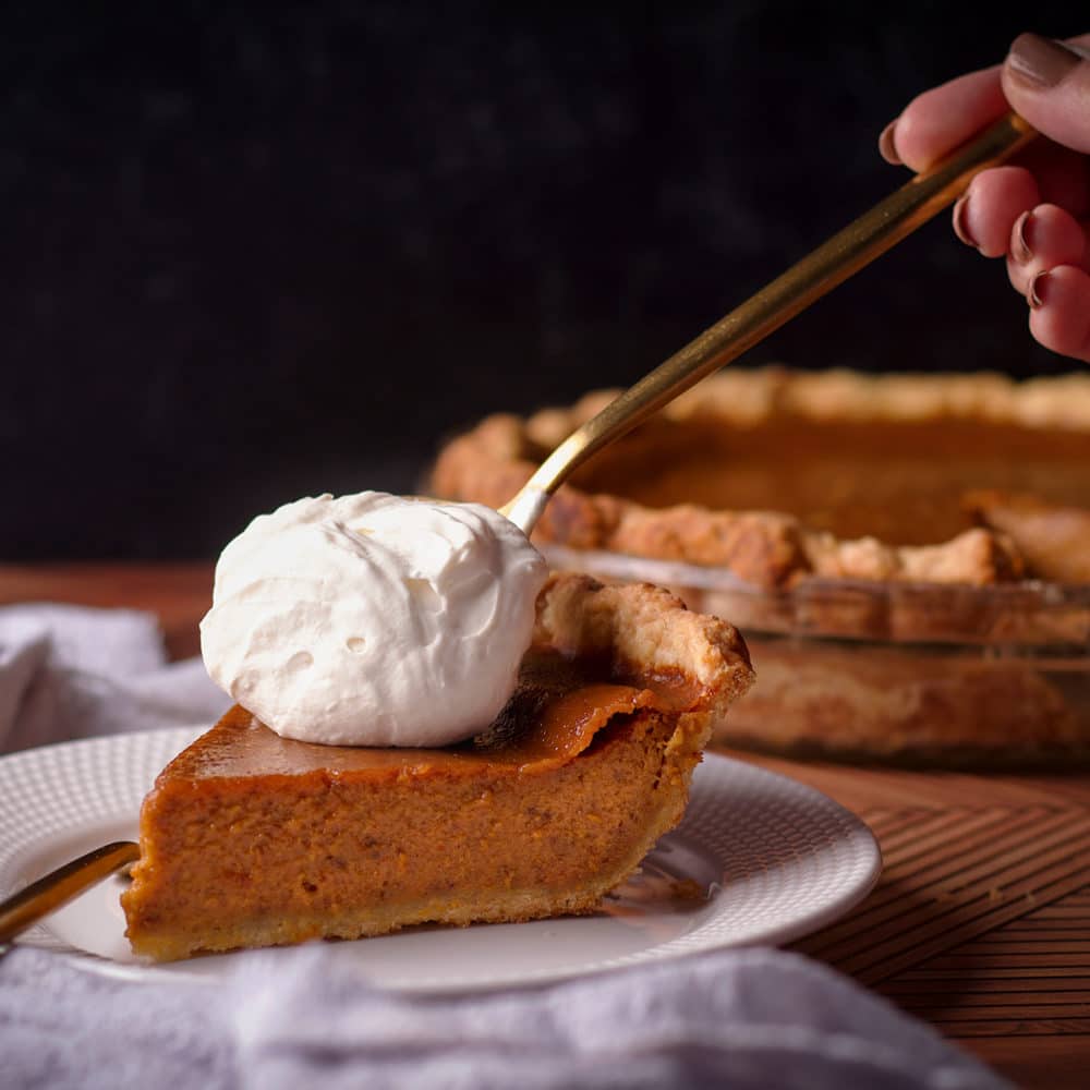 Someone using a gold spoon to top a slice of Maple Pumpkin Pie with a dollop of Maple Whipped Cream