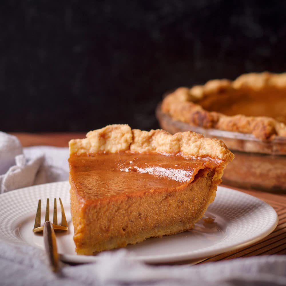 A slice of Maple Pumpkin Pie on a white plate with a gold fork and the entire pumpkin pie in the background.