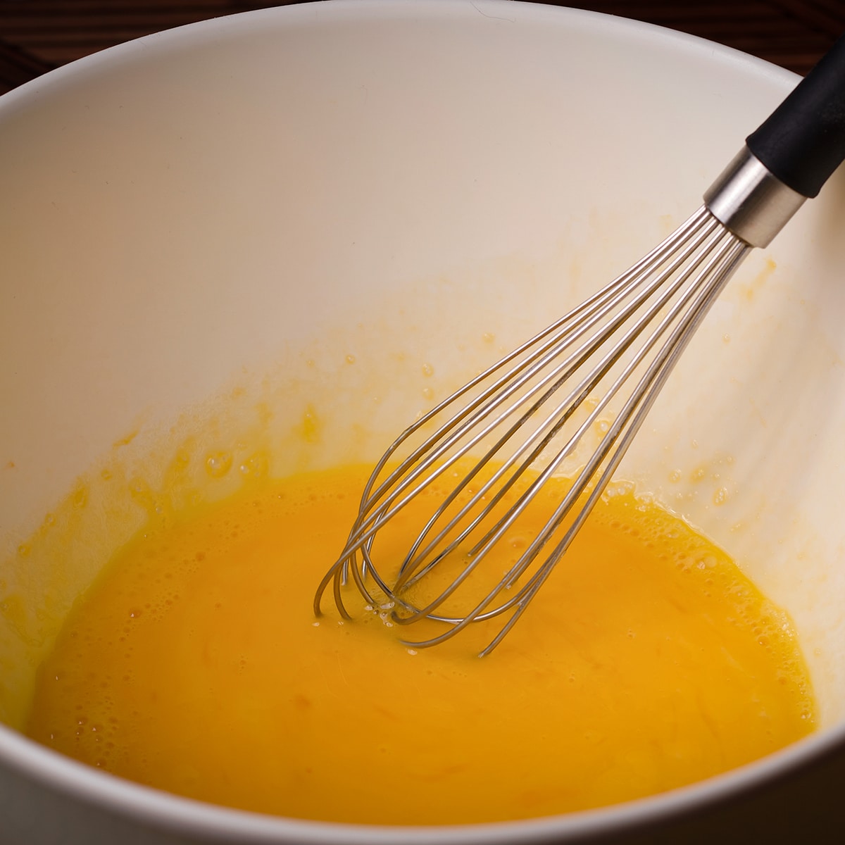 Someone using a wire whisk to beat eggs in a white bowl.
