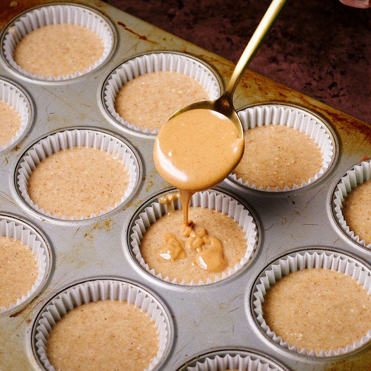 Someone using a gold spoon to drizzle melted peanut butter over gluten free banana muffin batter.