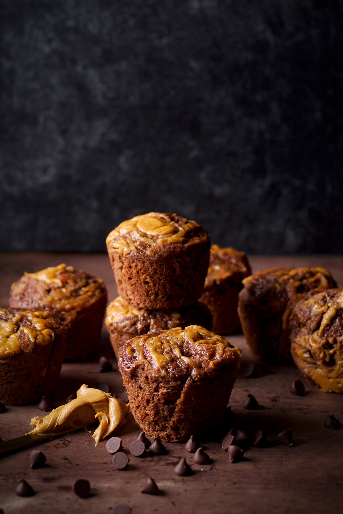Chocolate banana muffins with a peanut butter swirl stacked on a table with a peanut butter covered knife laying next to them.