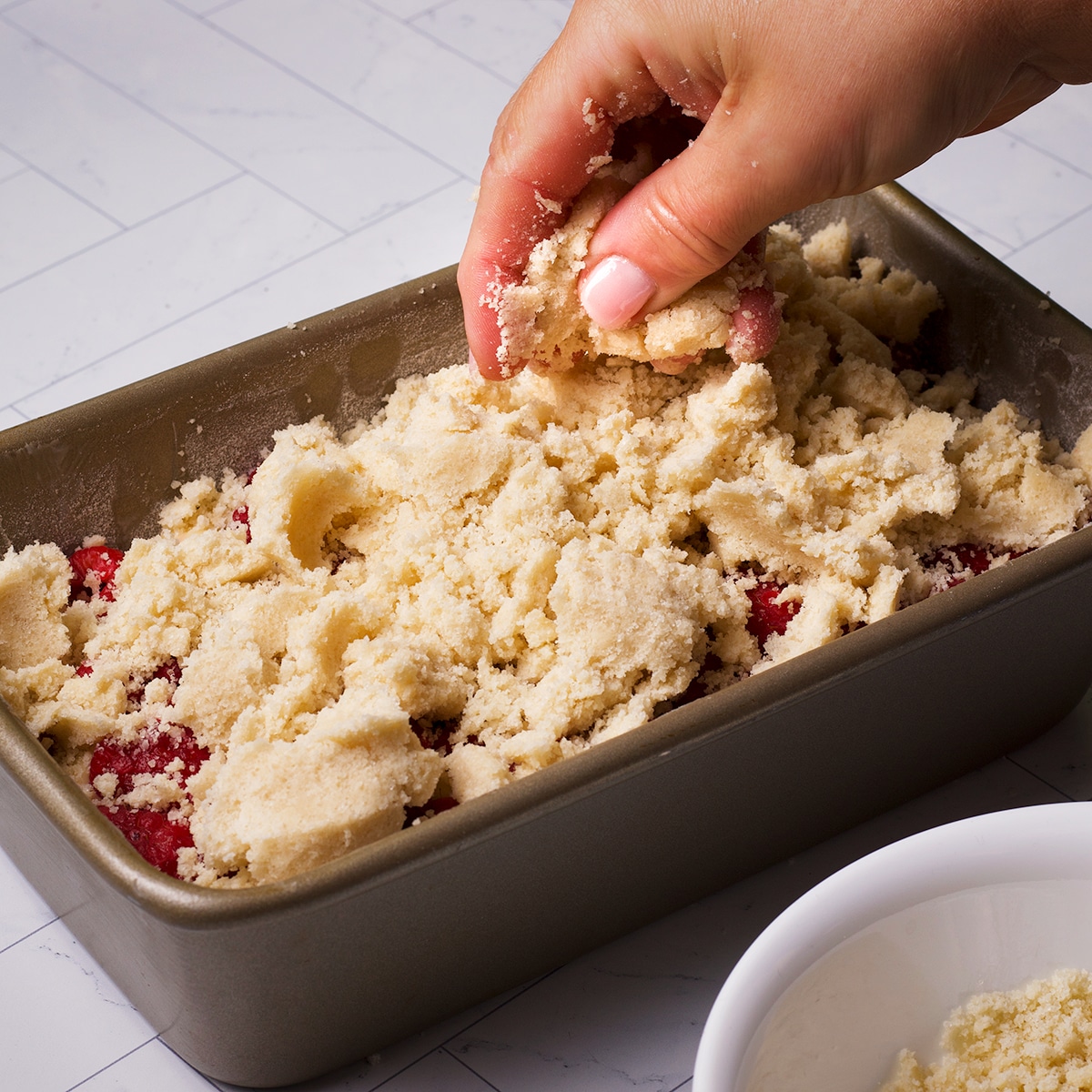 Someone using their fingers to squeeze the crumb topping together to create large crumbs as they top the raspberry bread batter with the topping.