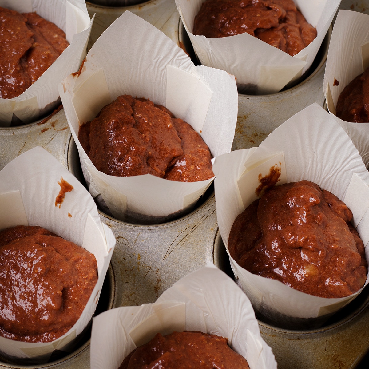 A muffin tin containing tulip paper liners that have been filled with chocolate banana muffin batter.
