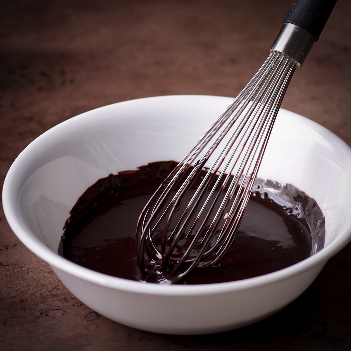 Someone using a wire whisk to mix up a simple chocolate glaze for double chocolate banana muffins.