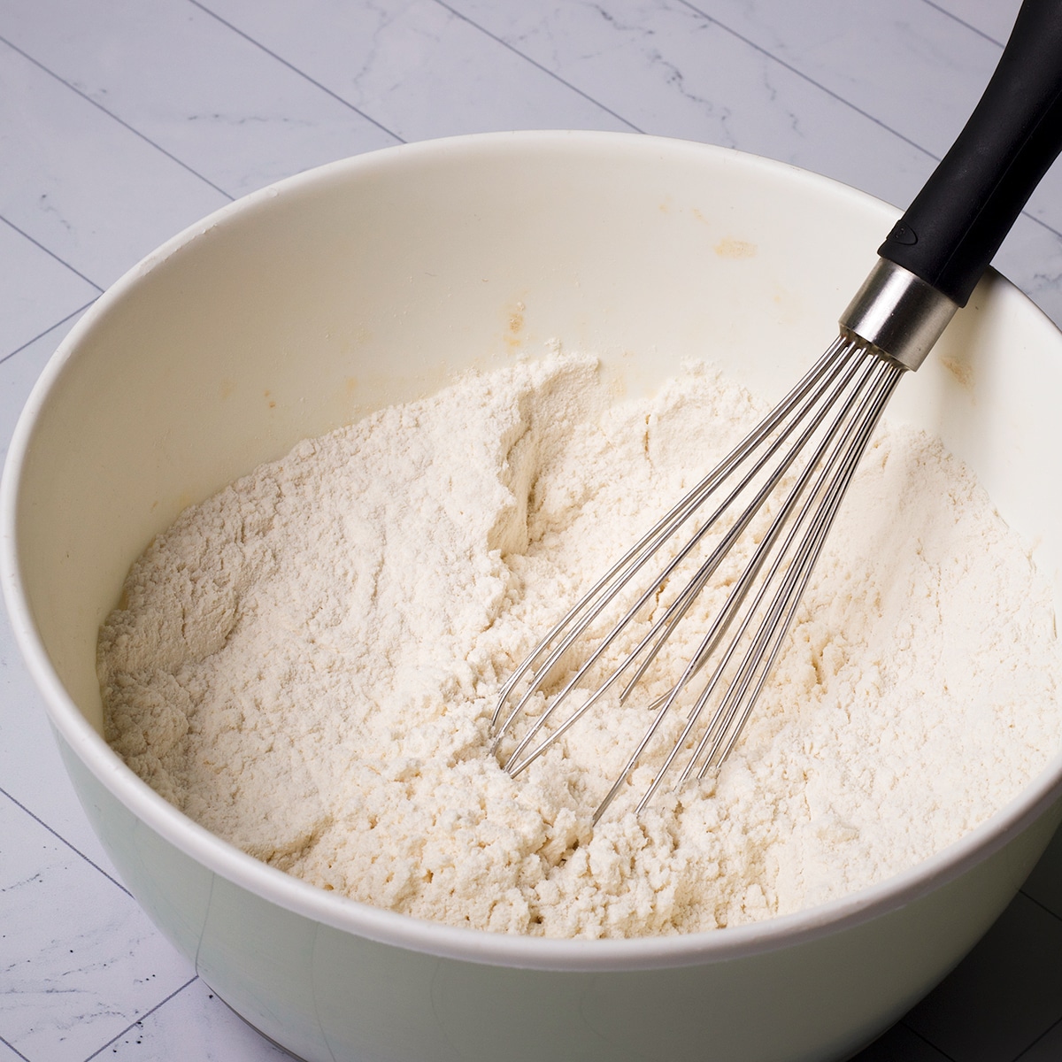 Using a wire whisk to stir flour, baking powder, baking soda, and salt in a white bowl.