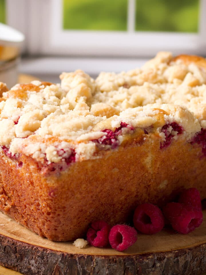 A loaf of raspberry bread with crumb topping on a wood serving board surrounded by fresh raspberries.
