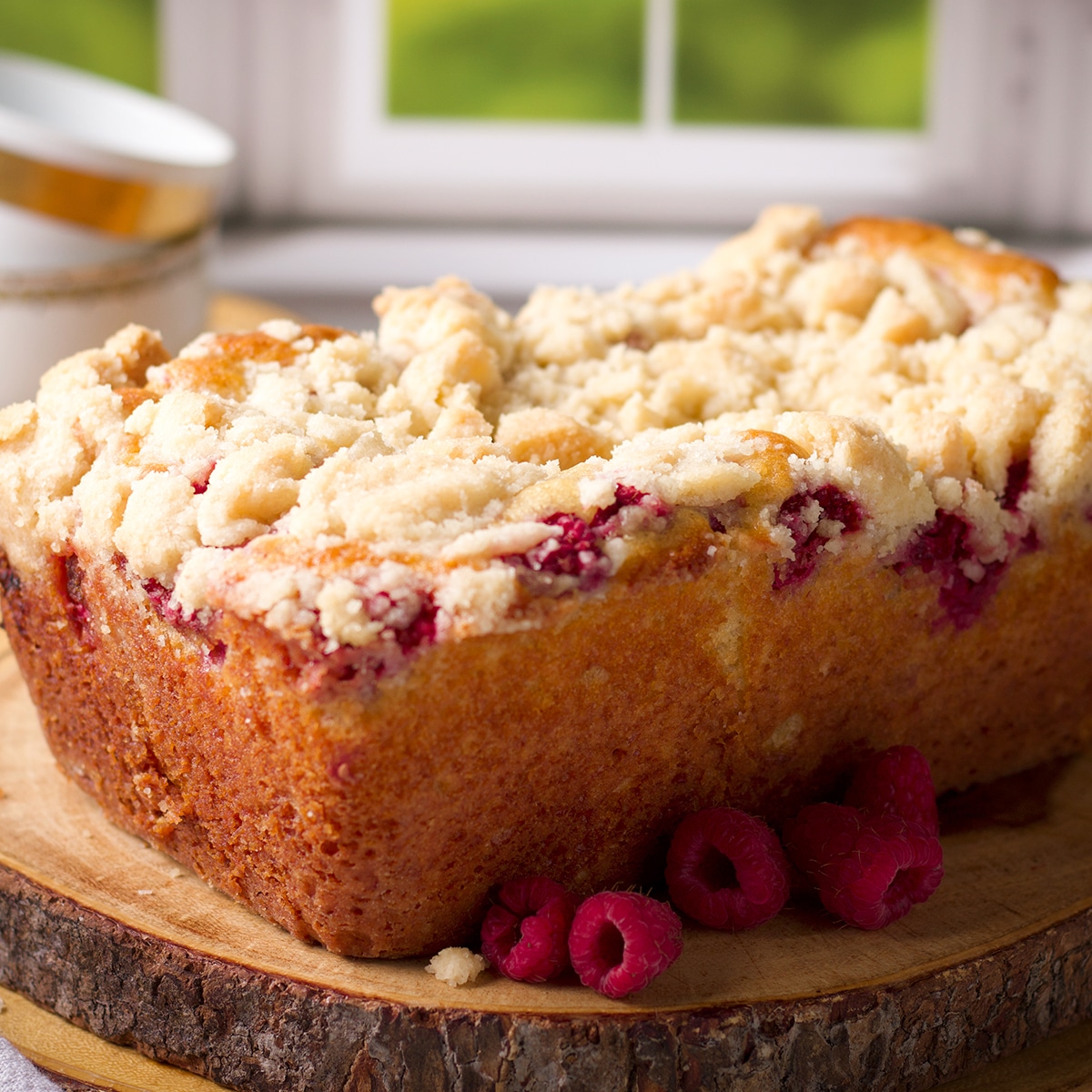 A loaf of raspberry bread with crumb topping on a wood serving board surrounded by fresh raspberries.