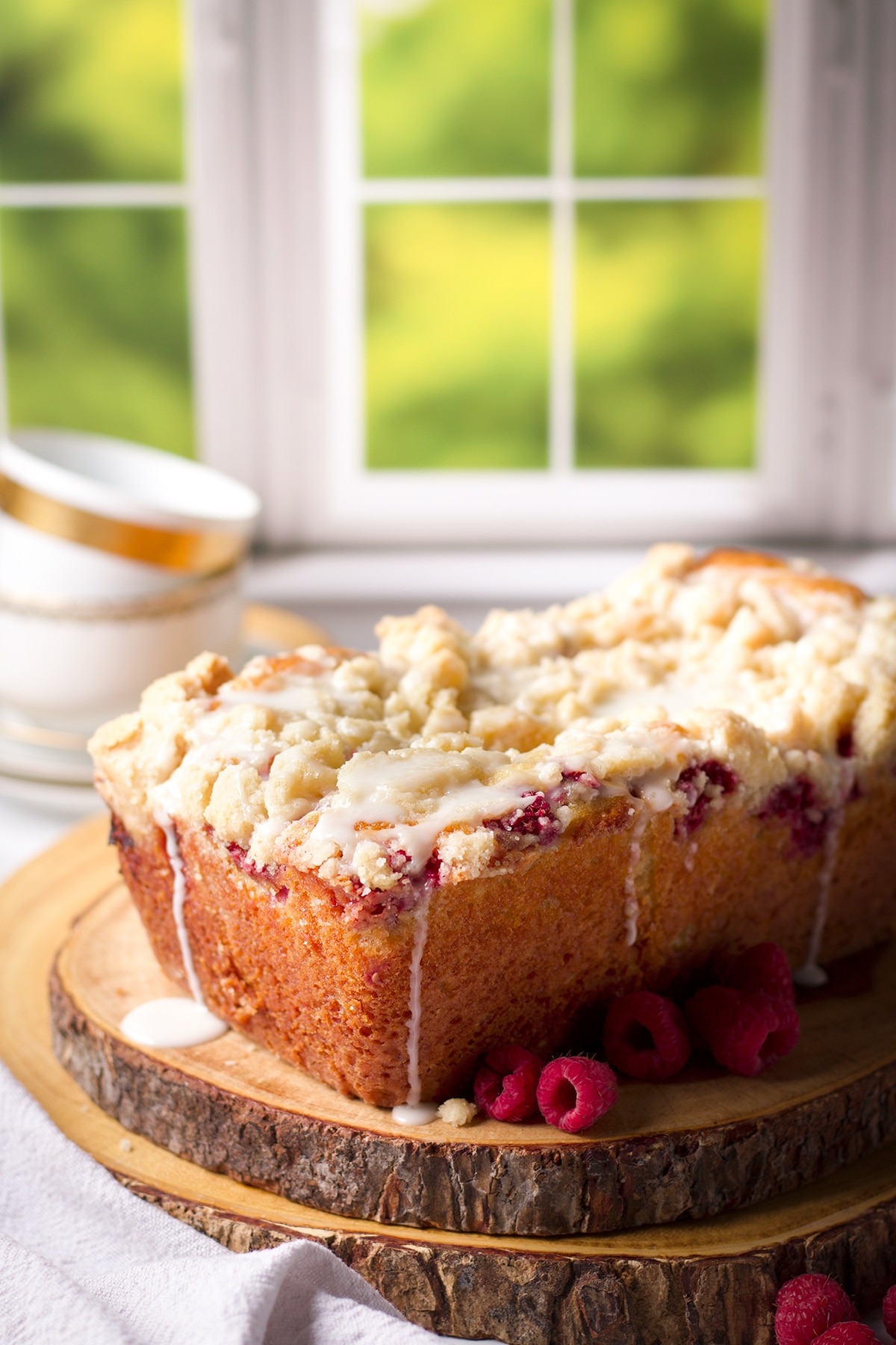 A loaf of raspberry bread topped with lemon icing on a wood cake board with fresh raspberries all around it.