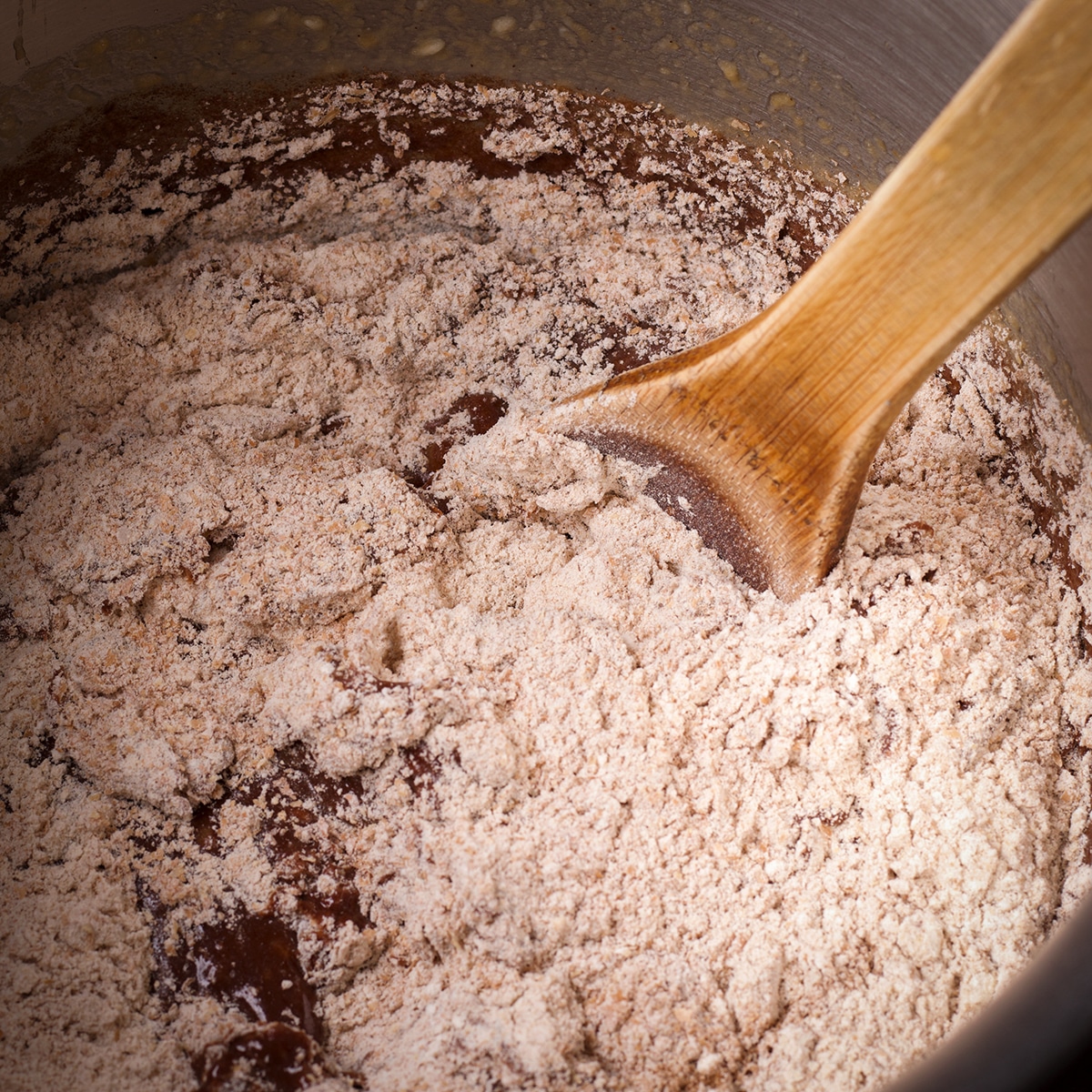 Someone using a wooden spoon to stir the dry ingredients into chocolate banana muffin batter.