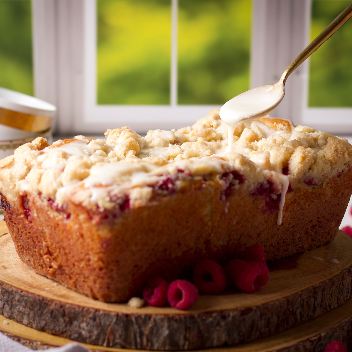 Using a spoon to drizzle lemon icing over the top of a loaf of raspberry bread with crumb topping.