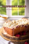 A loaf of raspberry bread topped with lemon icing on a wood cake board with fresh raspberries all around it.