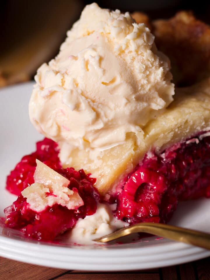 A slice of raspberry pie with a lattice crust topped with a scoop of vanilla ice cream.