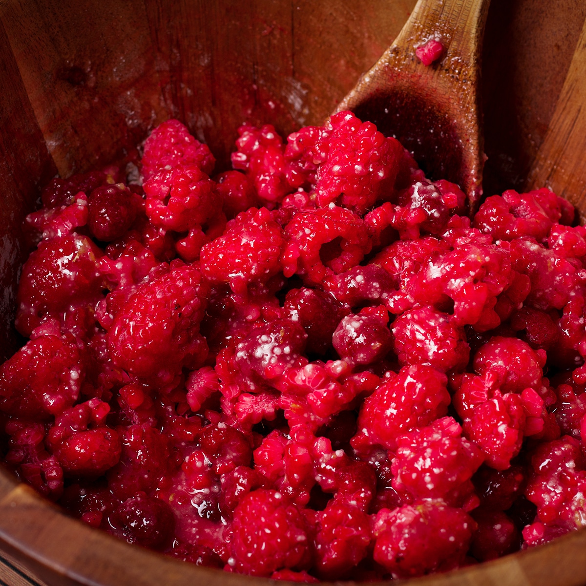 Using a wooden spoon to gently stir raspberries into the other ingredients for raspberry pie filling.