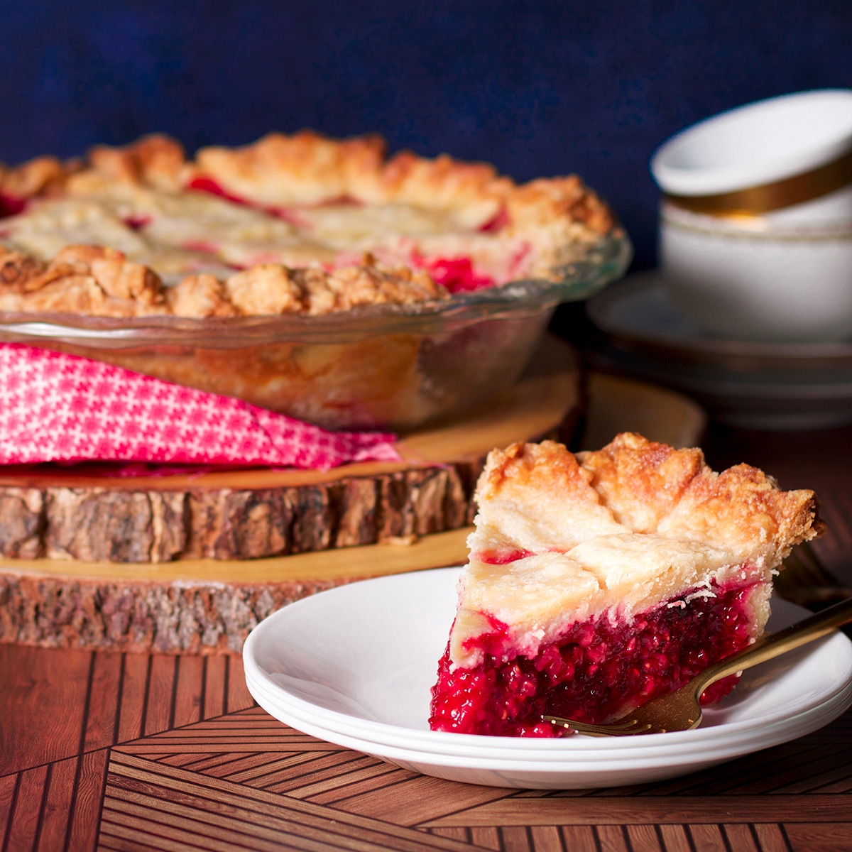 A slice of raspberry pie on a white plate sitting on a wood table next to a coffee cup.