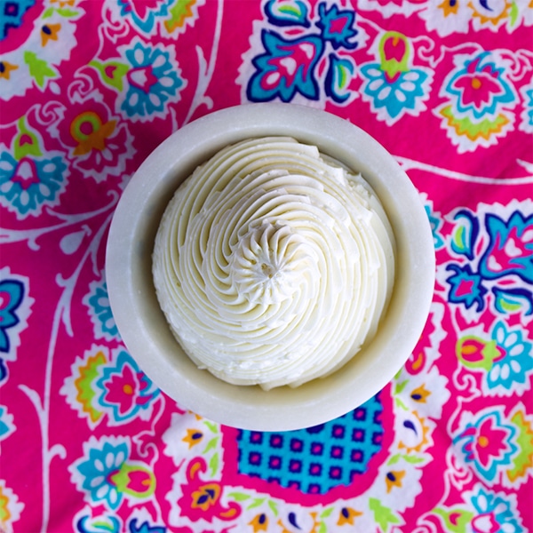 A small bowl filled with Italian Meringue Buttercream that's been piped into the bowl in a swirl. The bowl is sitting on a bright pink paisley tablecloth.