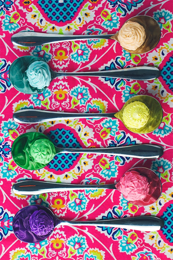 Six spoon laying side by side on a bright pink tablecloth. Each spoon holds a different color of Italian Meringue Buttercream. 