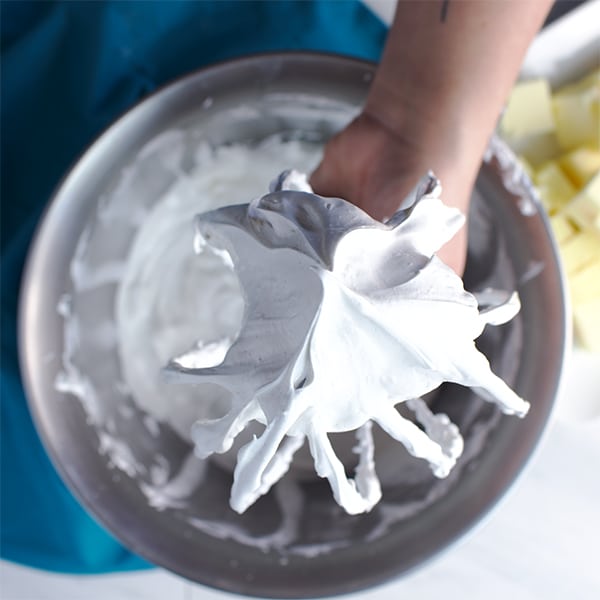 Someone holding up a beater that's been lifted from the bowl of a stand mixer to show meringue that's been beaten to the point of being able to hold a stiff peak.