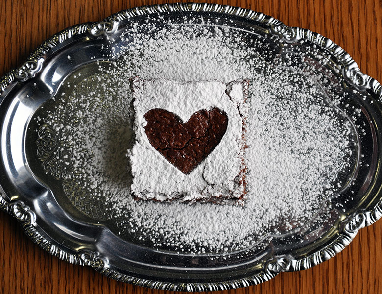 A dark chocolate brownie on a small silver tray. The brownie has been dusted with powdered sugar to create a heart shape on the top of the brownie.