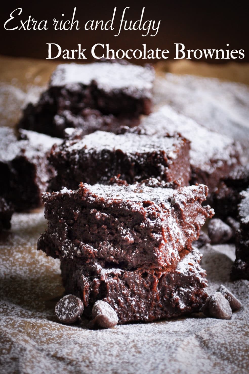 Two dark chocolate fudge brownies stacked on top of each other with more brownies in the background.