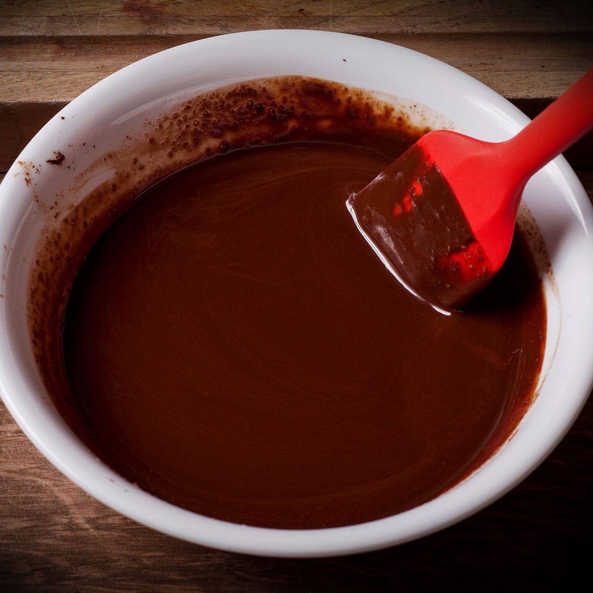 Using a red spatula to stir melted chocolate with melted butter in a white bowl.