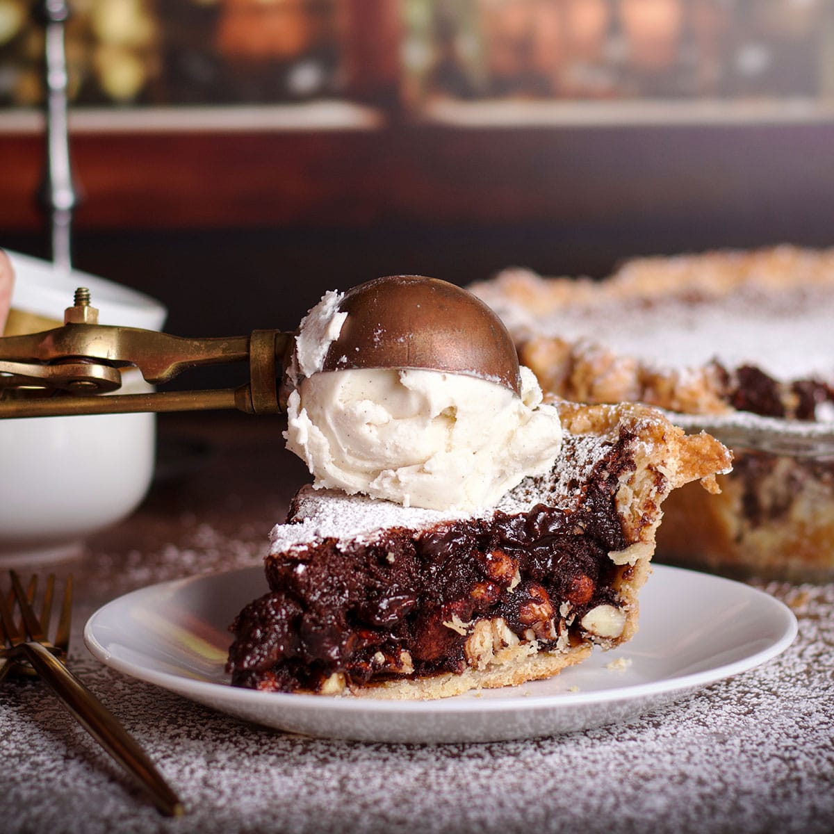 Someone using an old fashioned ice cream scoop to top a slice of chocolate almond pie with a scoop of vanilla ice cream.