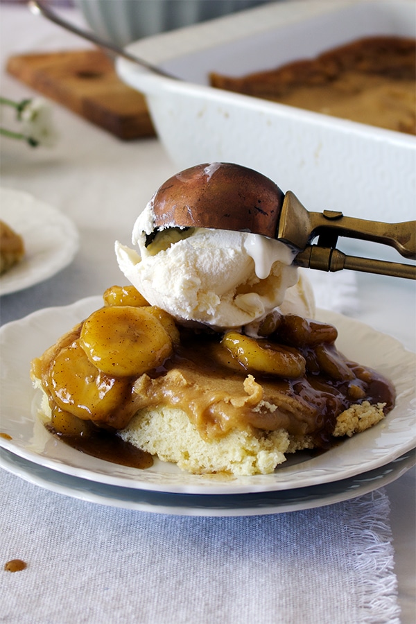 Someone using an ice cream scoop to top a piece of Bananas Foster Cake with vanilla ice cream. 