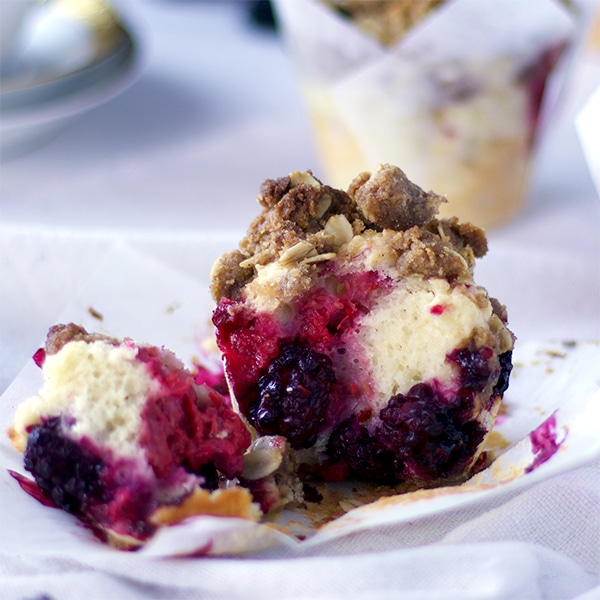 A mixed berry muffin with streusel on a table cover with a white tablecloth. The muffin has been broken in half so you can see the berry filled inside.