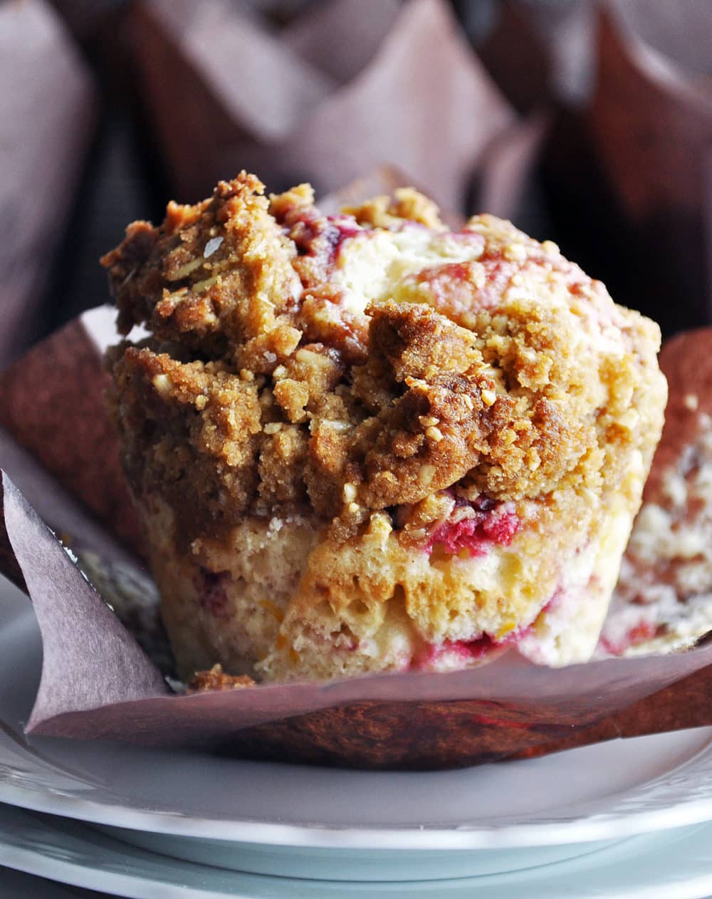 A berry muffin topped with streusel sitting on a table with other muffins in the background. The muffin's paper liner has been pulled back so you can see the entire muffin.