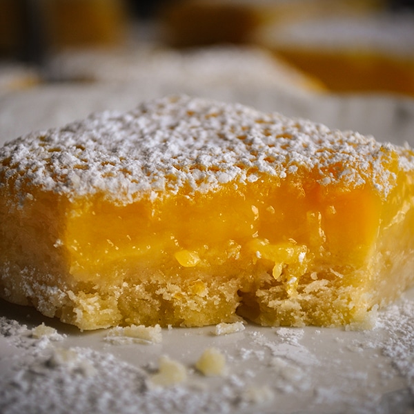 A lemon bar on a plate with a bite taken out of it so you can see the buttery shortbread crust and the creamy lemon filling.