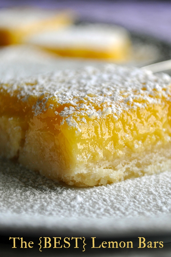 A lemon bar with a shortbread crust on a white plate with other lemon bars in the background. The square has been dusted with powdered sugar.