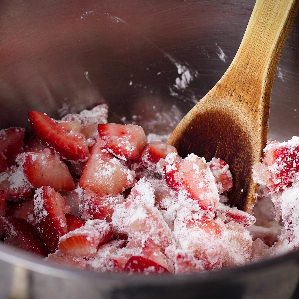 Someone using a wooden spoon to stir the ingredients for strawberry pie filling in a large saucepan.