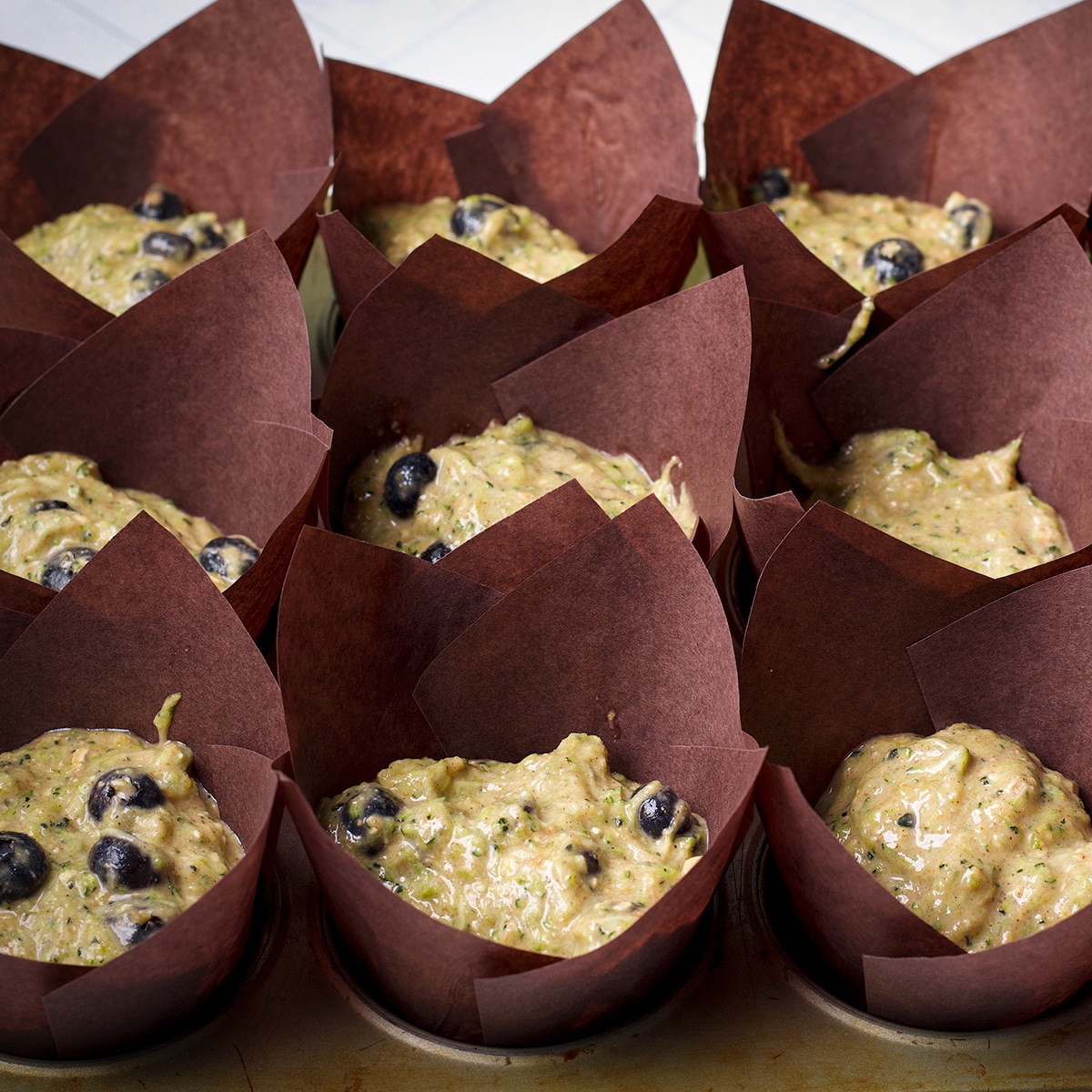 A muffin tray lined with tulip baking cups that are filled with zucchini blueberry muffin batter.