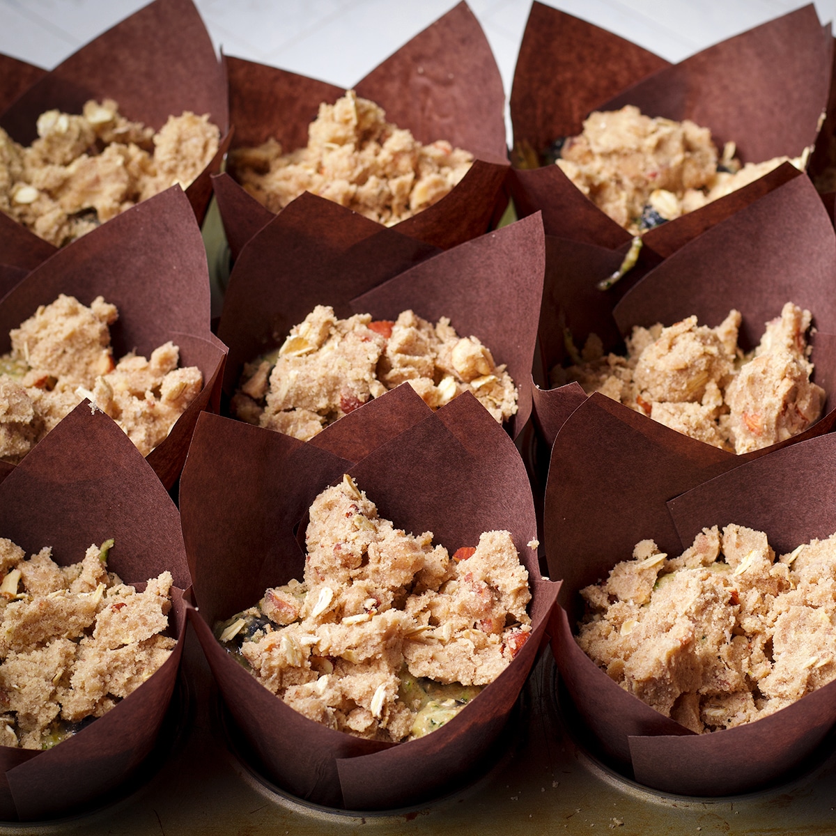A muffin tray lined with tulip baking cups that are filled with zucchini blueberry muffin batter topped with oat and almond streusel. 