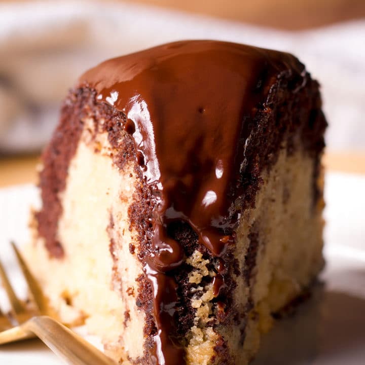 A slice of marble cake with chocolate glaze on a white plate with a gold fork.
