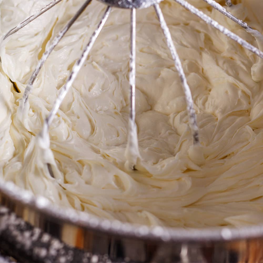 An electric stand mixer fitted with the whisk attachment beating a bowl full of cream cheese buttercream.