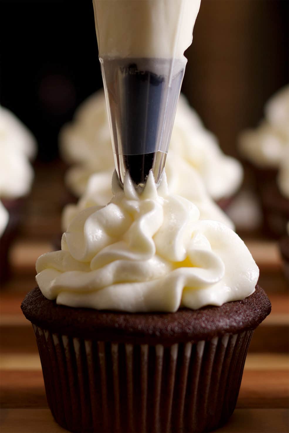 Someone piping cream cheese buttercream from a pastry bag onto the top of a chocolate cupcake.