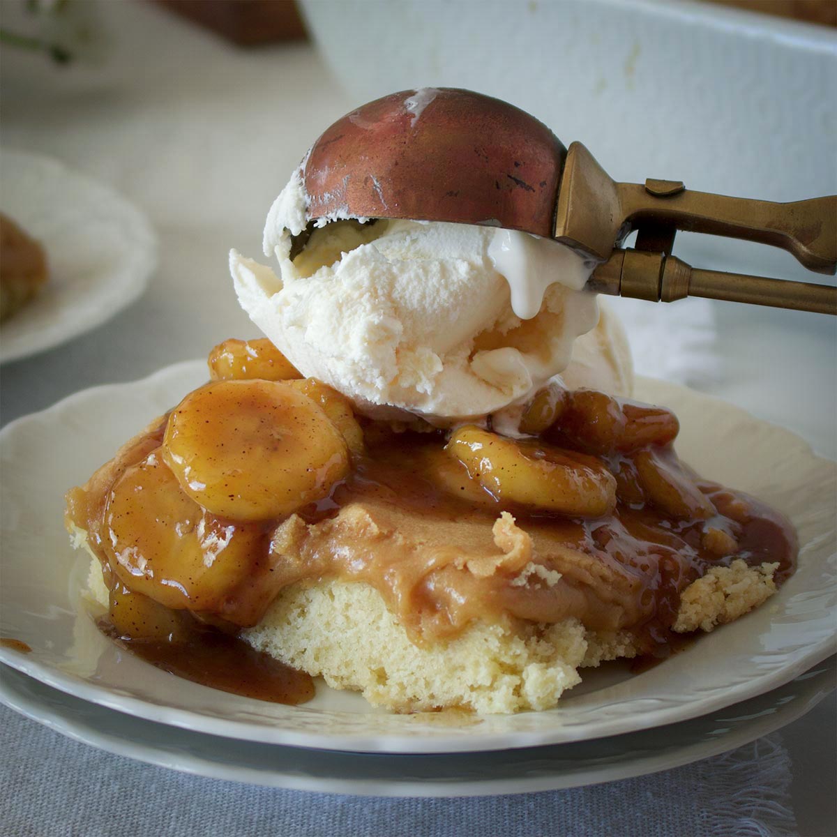 Someone using an old fashioned metal ice cream scoop to top a slice of bananas foster butter cake with vanilla ice cream.