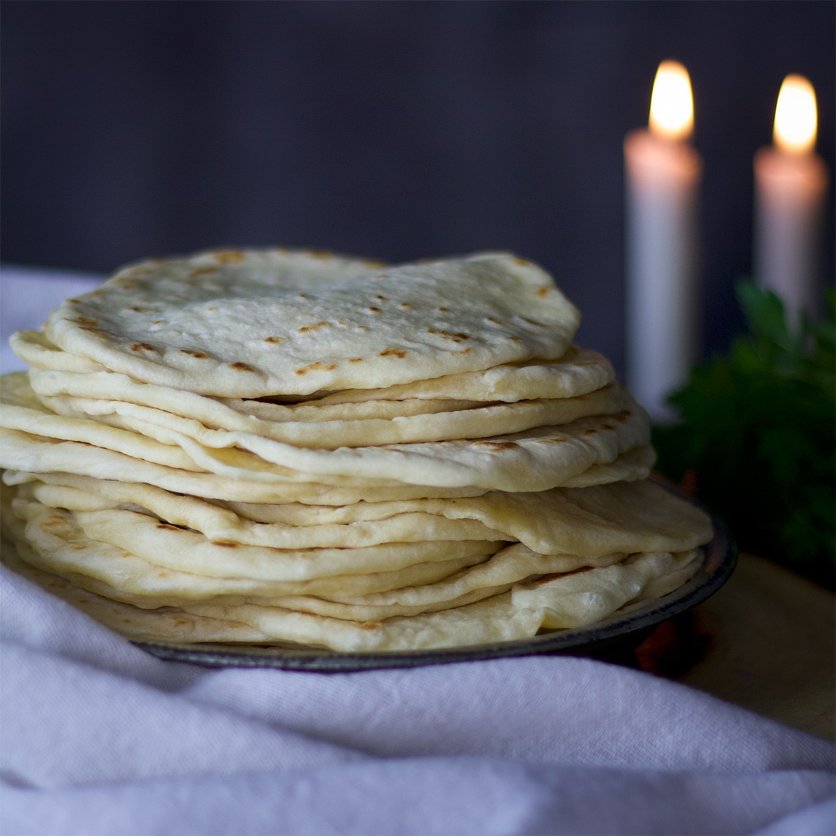 A tall stack of homemade flour tortillas on a table with lit candles behind them.
