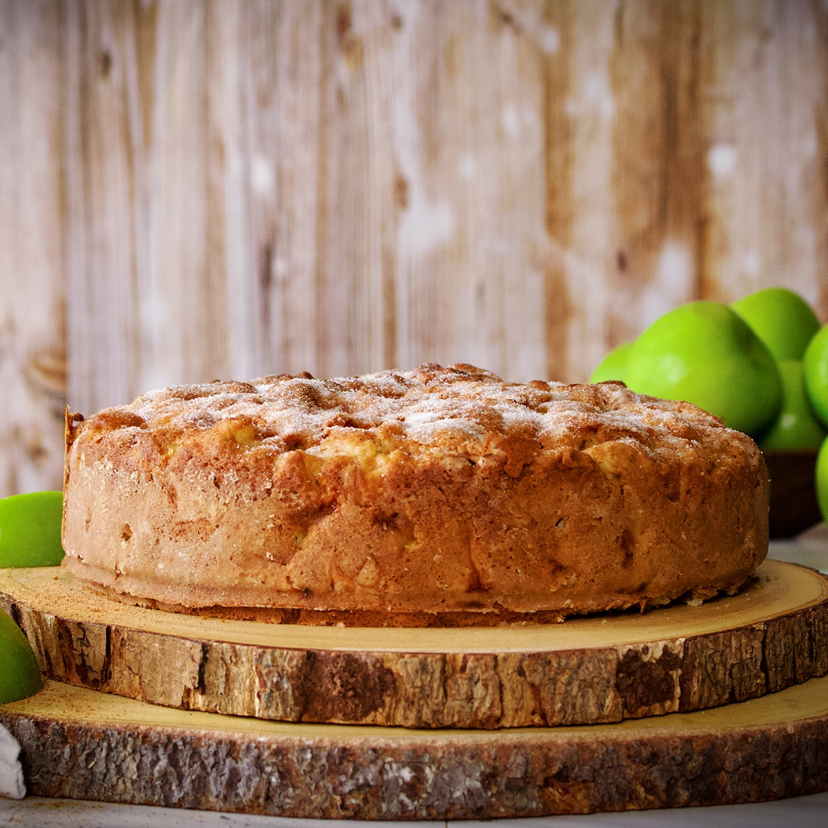 An Irish Apple Cake with a sugar crusted top on two wood cake boards surrounded by green apples.