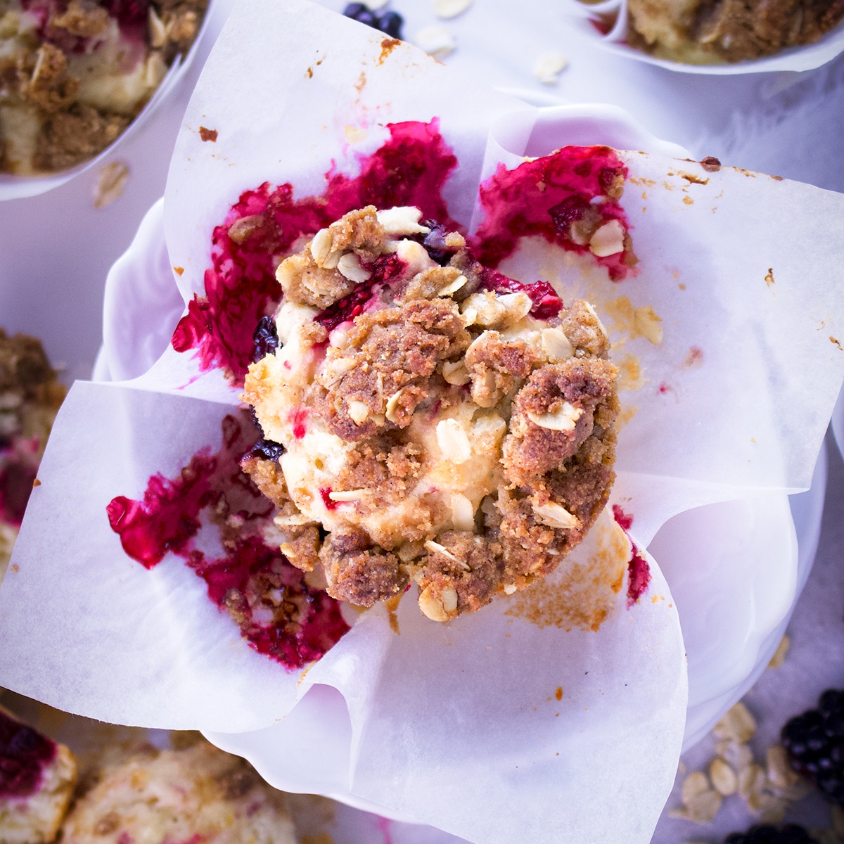 A mixed berry muffin with brown sugar and oats streusel in the center of a table covered in muffins and berries.