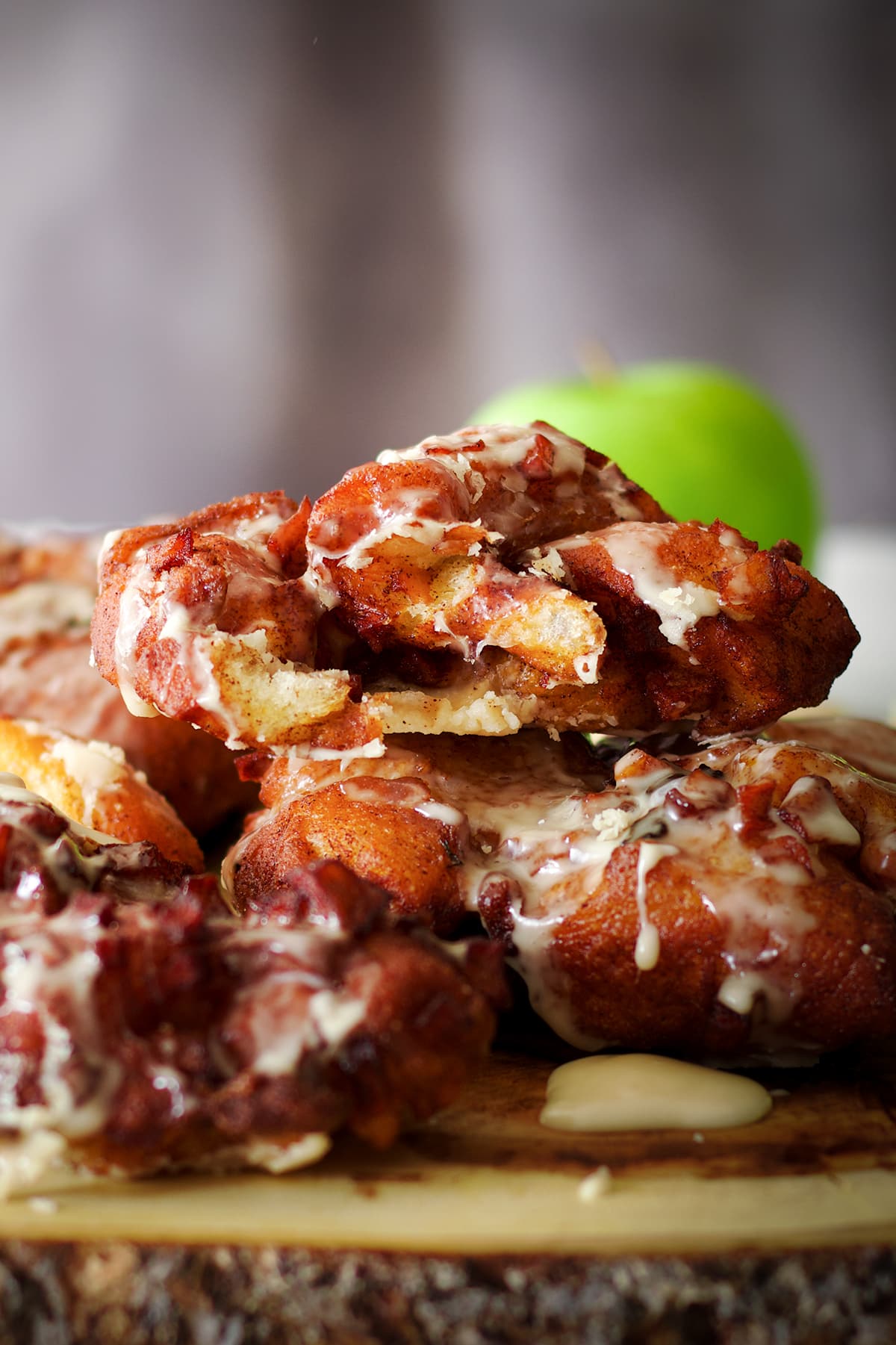 A stack of homemade apple fritters covered in maple glaze on a wood platter with green apples in the background.