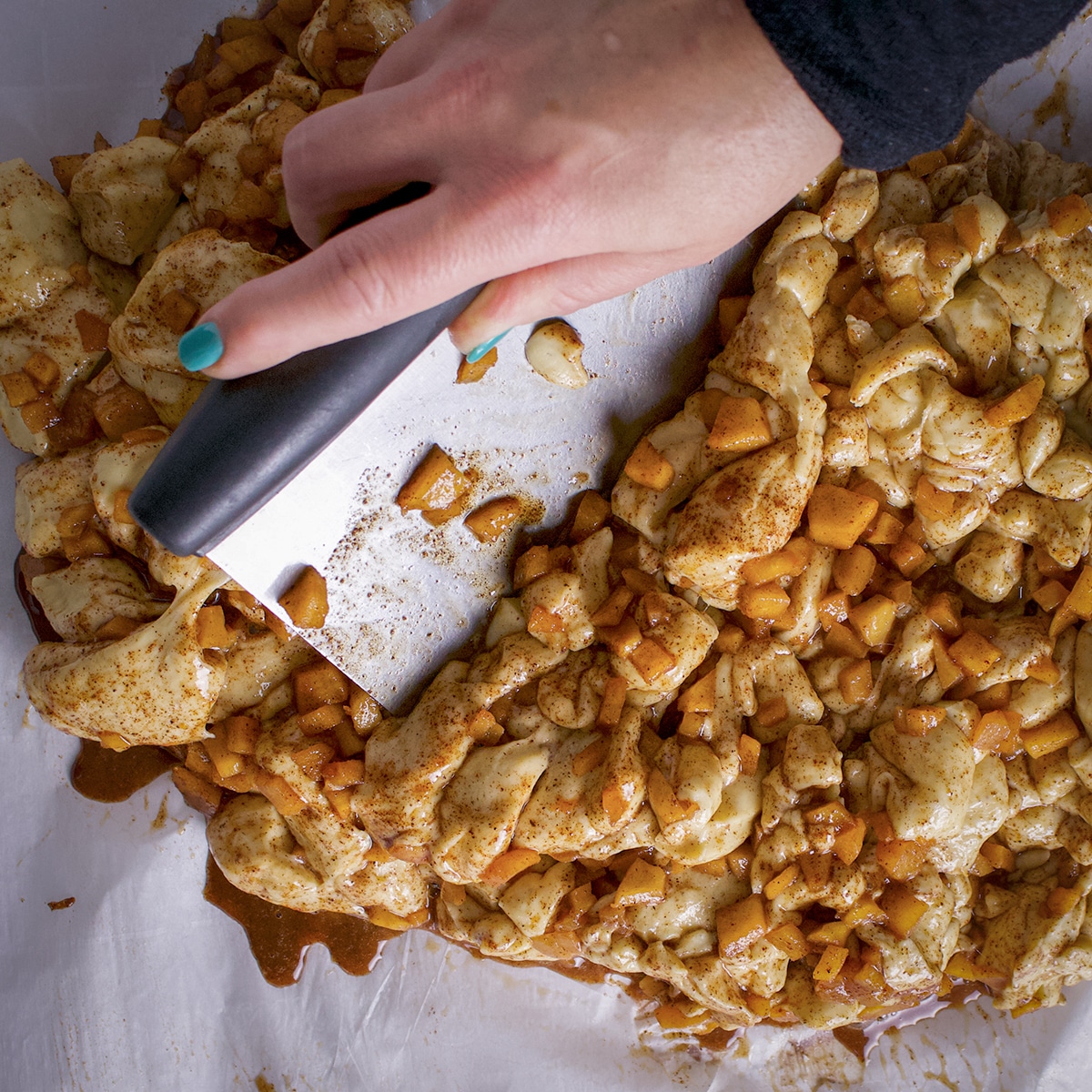 Someone using a dough scraper to chop pieces of apple fritter dough into many small pieces.