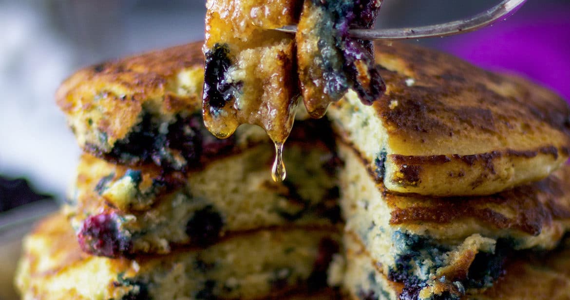 A bite of blueberry pancakes with maple syrup dripping from the fork.