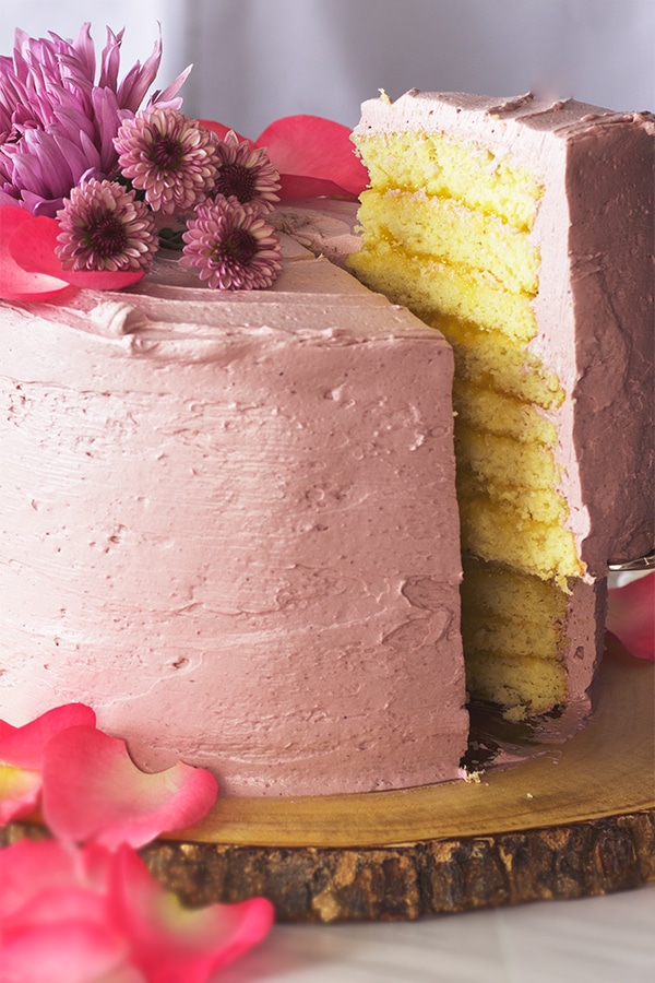 Someone using a cake server to lift a slice of Lemon Layer Cake covered in Blackberry Buttercream from the whole cake.
