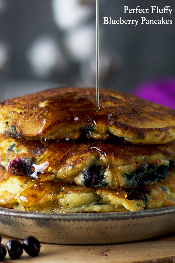 Pouring maple syrup over a plate of fluffy blueberry pancakes.