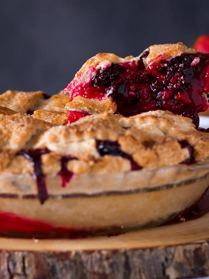 Someone using a pie server to lift a slice of triple berry plum pie from the whole pie.