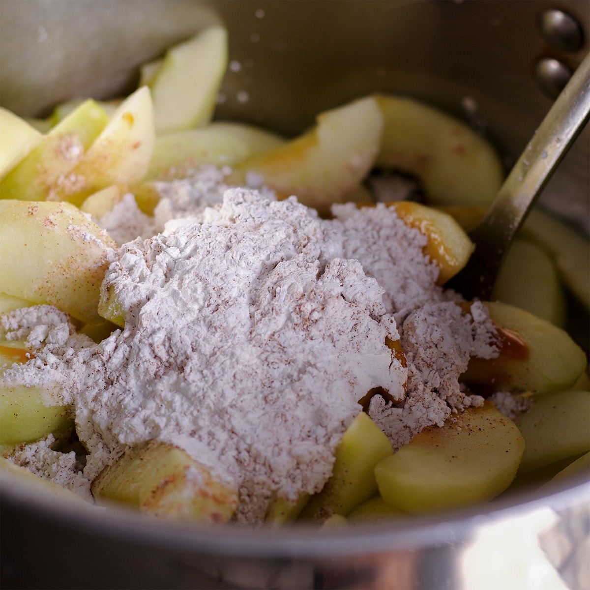 Someone using a spoon to stir flour into a saucepan of sliced apples and sugar.