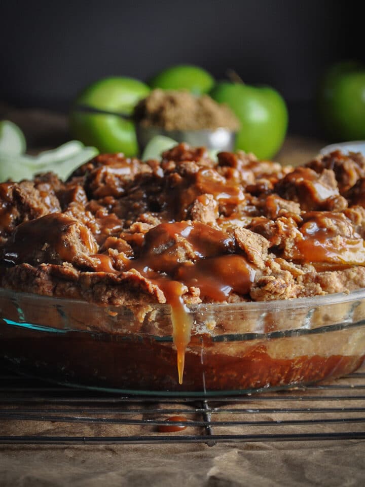 A salted caramel apple pie with crumb topping cooling on a wire rack. Caramel sauce is dripping over the side of the pie plate.