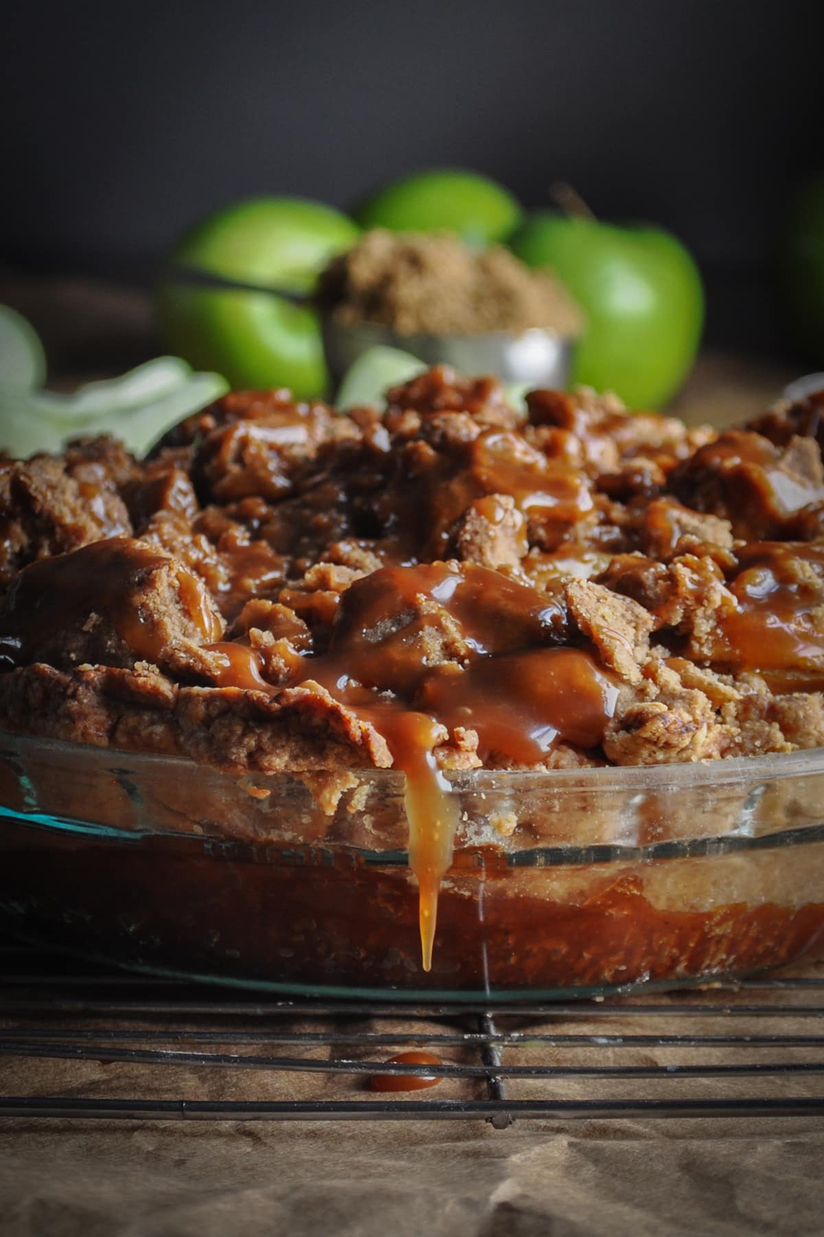 A salted caramel apple pie with crumb topping cooling on a wire rack. Caramel sauce is dripping over the side of the pie plate.